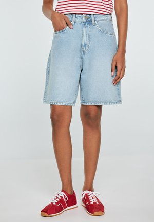 Osoba ubrana w jasnobłękitne szorty z denimu, czerwono-biały pasiasty top oraz czerwone trampki z białymi sznurowadłami, stojąca z jedną ręką w kieszeni.