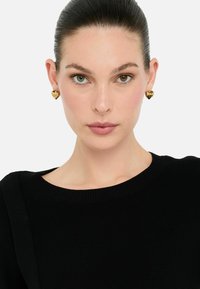 Orecchini a forma di cuore in oro con dettagli in rilievo. Il modello indossa un maglione nero con una texture a coste e un ampio scollo.