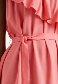 Korálové šaty s volány na výstřihu, staženým pasem s látkovým páskem, uvázaným do mašle, a měkkou, splývavou texturou látky.