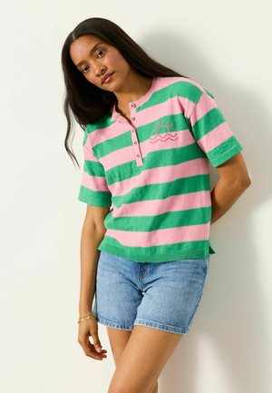 Frau mit langen dunklen Haaren, die ein grün-rosa gestreiftes kurzärmeliges Shirt mit Sonnenstickerei und blaue Jeansshorts trägt, steht vor einer weißen Wand.