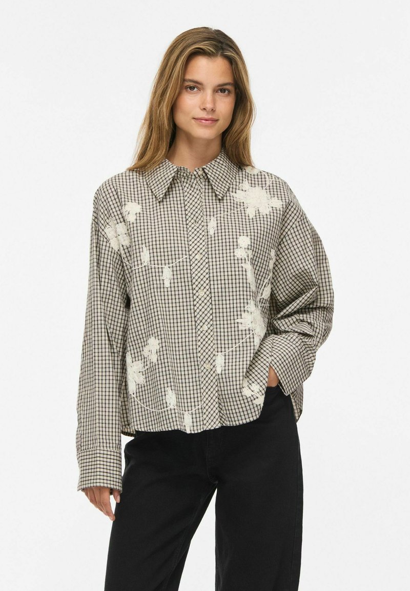 Femme aux longs cheveux portant une chemise oversize à carreaux beige et noire avec des imprimés floraux blancs et un pantalon noir, posant avec une main dans la poche.