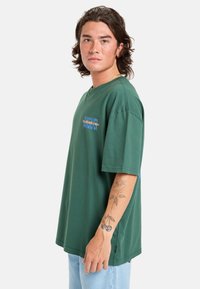 Camiseta de algodón verde con mangas cortas, que presenta un logo en azul y naranja en el frontal y un ajuste casual y holgado.