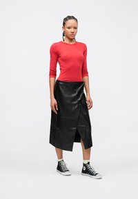 Jonge vrouw met gevlochten haar, gekleed in een rood shirt met lange mouwen, een zwarte leren midirok met een vooraan gespleten stuk, witte sokken en zwarte hoge sneakers.