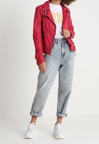 Czerwona skórzana kurtka motocyklowa z detalami na suwakach, noszona na białej koszulce z żółtym napisem, zestawiona z jasnoniebieskimi, wysokimi spodniami dżinsowymi i białymi trampkami.