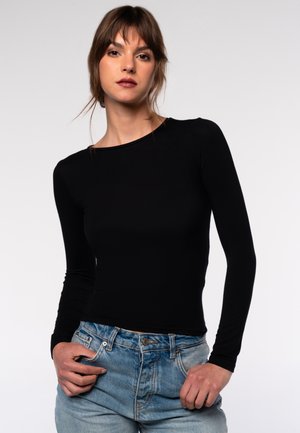 Schwarzes, langärmliges, tailliertes Oberteil aus dehnbarem Material, kombiniert mit hellblauen, hochgeschnittenen Jeans. Einfaches Design, glatte Textur.