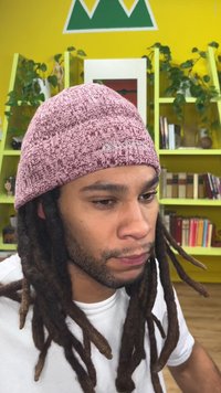 Ένα άτομο με σκούρα, πλεξούδες μαλλιών φοράει ένα υφασμάτινο ροζ σκουφάκι και ένα λευκό πουκάμισο, με βιβλιοθήκες και έναν κίτρινο τοίχο στο παρασκήνιο.