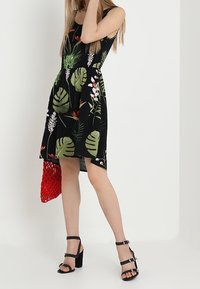 Svart klänning med färgglad tropisk blad- och blommiga tryck, utsvängd design, i kombination med en röd nätväska och svarta klacksandaler.