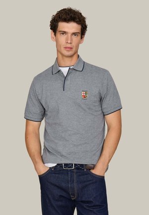 Hombre con camiseta tipo polo gris de manga corta con escudo bordado y vaqueros azul oscuro, de pie con las manos en los bolsillos contra un fondo liso.