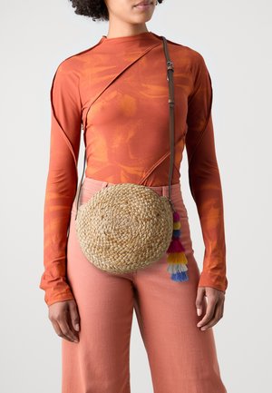 Nainen, joka pukeutunut ruosteenoranssiin pitkähihkaiseen yläosaan ja vaaleanpunaisiin housuihin, kantaa pyöreää, monivärisillä tupsuilla koristeltua punottua olkalaukkua.