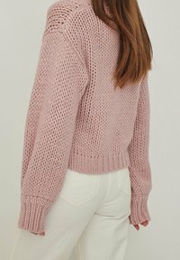 Gruby sweter z dzianiny w delikatnym różowym kolorze z prążkowanymi mankietami i dołem. Posiada luźny krój i fakturowany wzór, zestawiony z białymi spodniami z denimu.