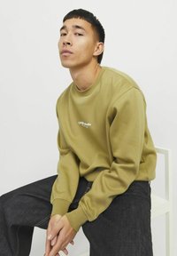 Olijfgroen sweatshirt met een ronde hals en geribbelde manchetten, voorzien van een wit logo op de borst. Gepaard met een donkere spijkerbroek.