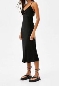 Svart satin spetklänning med en rosett vid halslinjen, tunna remmar och en flytande siluett, tillsammans med svarta sandaler med remmar.