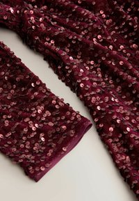 Burgunderfarbener, mit Pailletten besetzter Stoff mit einer weichen Textur, ausgestattet mit eng zusammenliegenden, runden Pailletten. Die Ärmel sind mit Bündchen versehen, was dem Design zusätzliche Details verleiht.