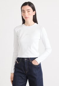 Il modello posa indossando una camicia bianca a maniche lunghe con un logo, abbinata a jeans scuri, su uno sfondo neutro. La luce naturale mette in risalto i suoi tratti.