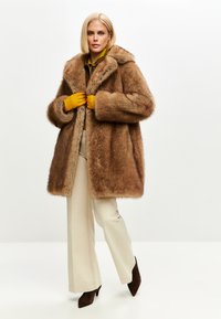 Brun faux fur-frakke med en bred krave, enkeltknaplukning og lange ærmer. Komplementeres af beige vidde bukser og brune hæle.