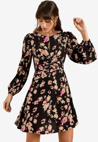Rochie neagră cu flori, având un față răsucită, mâneci voluminoase lungi și o fustă evazată. Culorile includ modele florale în nuanțe de roz, bej și crem.