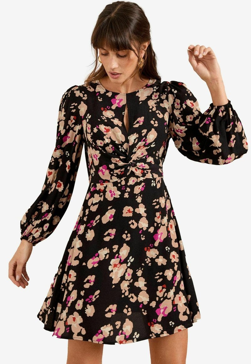 Rochie neagră cu flori, având un față răsucită, mâneci voluminoase lungi și o fustă evazată. Culorile includ modele florale în nuanțe de roz, bej și crem.