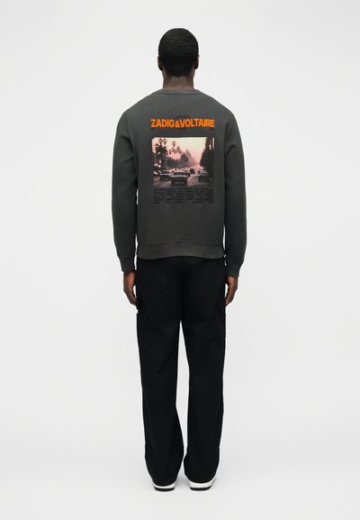 Άνδρας που φαίνεται από πίσω φοράει σκούρο φούτερ με πορτοκαλί κείμενο "ZADIG & VOLTAIRE" και μια αστική σκηνή με αυτοκίνητα και φοίνικες στην πλάτη.