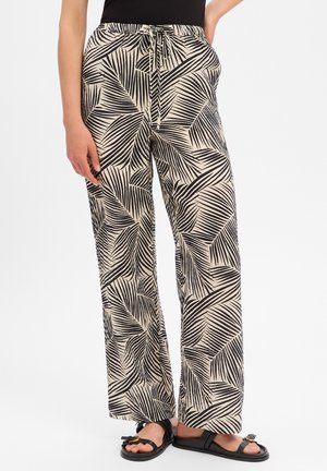 Pantalon ample beige avec motif de feuilles de palmier noires, taille à cordon, porté avec des sandales noires et un haut noir sur fond blanc.