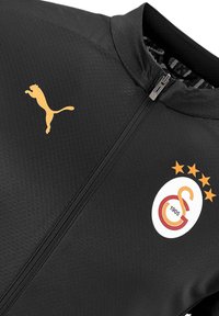 Schwarze Sportjacke mit Reißverschluss, versehen mit einem goldenen Puma-Logo und einem runden Logo mit orangefarbenen Akzenten und fünf Sternen.