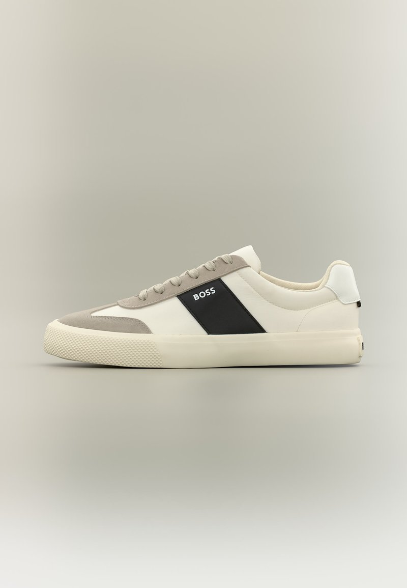Baskets basses blanches et gris clair avec panneau latéral noir arborant le logo "BOSS", semelle en caoutchouc texturée et lacets beiges sur fond neutre.