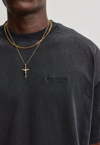 Dark-skinned person wearing a black shirt with "Pecador" logo and two gold necklaces, one with a crucifix pendant.