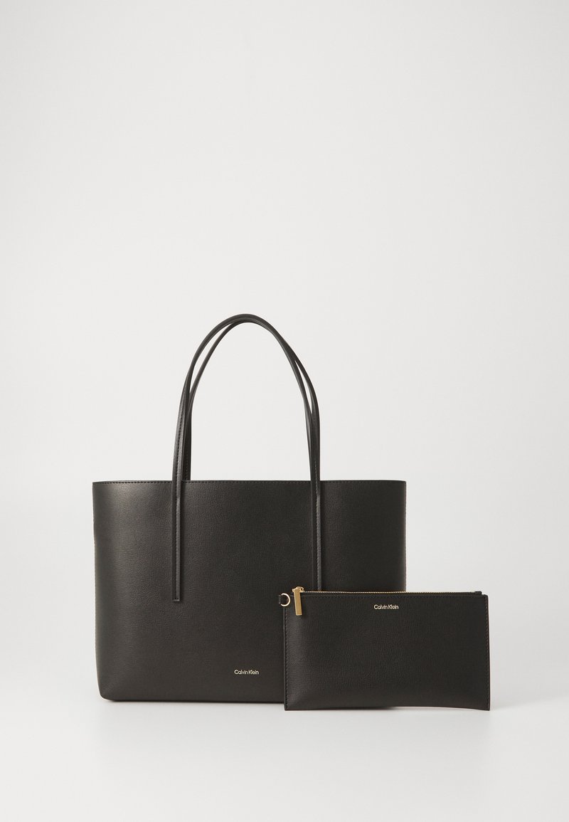 Svart tote bag med to håndtak, kombinert med en matchende glidelås-pung. Begge har et Calvin Klein-logo preget i sølv.