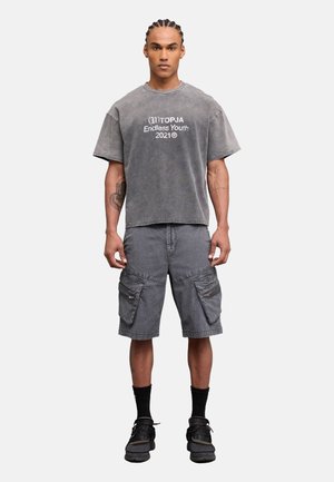 Hombre con cabello trenzado que lleva una camiseta gris extragrande con texto blanco, pantalones cortos cargo grises, calcetines negros y zapatillas negras, de pie.