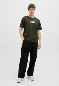 Mørkegrøn bomulds T-shirt med et hvidt trykt logo. Sammen med løse sorte bukser og hvide sneakers med beige detaljer.