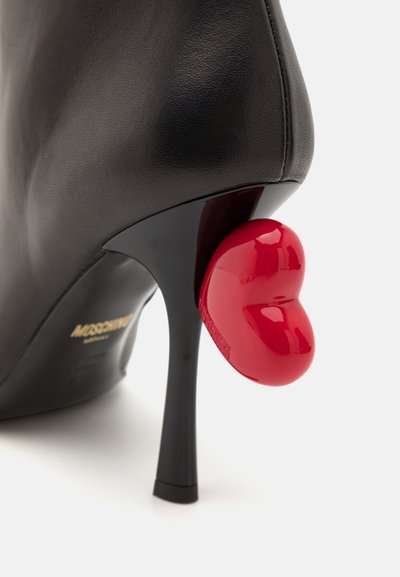 Black leather high-heeled shoe with a sleek design. Features a glossy red heart-shaped heel accent and a smooth texture. Gold branding visible.