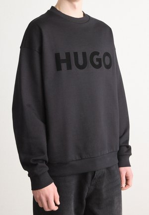 Person iført en sort oversize sweatshirt med "HUGO" trykt i store sorte bogstaver på tværs af brystet, kombineret med mørke bukser.