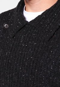 Černá pletená bunda s texturovaným vzorem, širokým límcem a výrazným knoflíkem. Materiál vypadá jako vlněný s jemnými barevnými skvrnami.