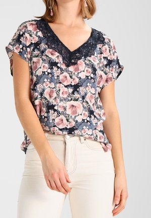 Frau trägt eine kurzärmelige geblümte Bluse mit schwarzem Spitzen-V-Ausschnitt und hellen Hosen vor einfarbigem Hintergrund.