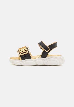 Sort og guld sandal med tyk hvid sål og justerbar rem, med guldskrift på den forreste rem.
