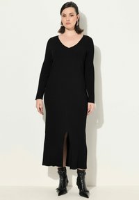 Schwarzes, geripptes Strickkleid mit langen Ärmeln und V-Ausschnitt. Es hat einen Schlitz vorne und eine Midi-Länge bis zur Wadenmitte. Kombiniert mit schwarzen Ankle Boots.