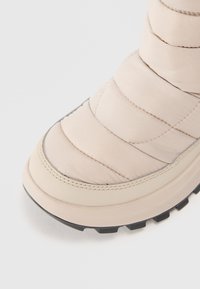 Stivale imbottito di un beige chiaro con una texture liscia, suola spessa e suola in gomma nera. Presenta cuciture orizzontali e punta rotonda.