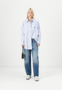 Striped button-up shirt in light blue and white, paired with loose blue jeans and gray shoes. Small pocket logo detail. Black bag accessory.