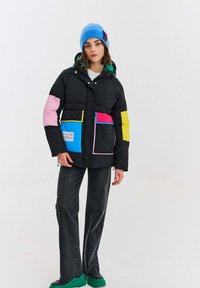 Žena v čiernej puffovej bunde s farebnými nášivkami, modrom čiapke, čiernych džínsach a zelených robustných topánkach, stojí pred bielym pozadím.