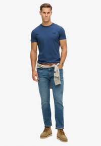 Marineblauwe t-shirt met korte mouwen, lichte spijkerbroek, bruine riem, tan laarzen, grijze trui om de taille geknoopt. Heeft een gestructureerde stof en een eenvoudig ontwerp.