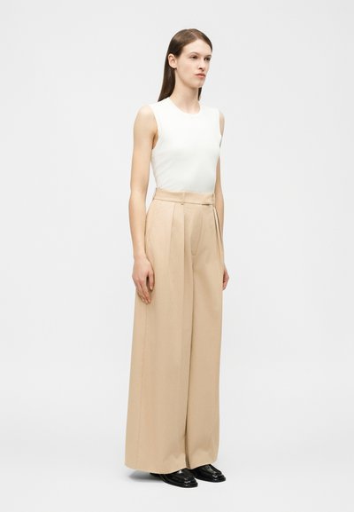 Woman standing wearing a white sleeveless top, beige high-waisted wide-leg trousers, and black shoes against a plain background.