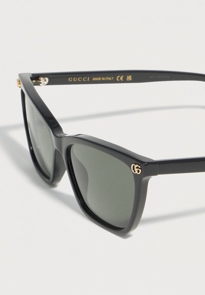 Μαύρα υπερμεγέθη τετράγωνα γυαλιά ηλίου Gucci με σκοτεινούς φακούς και χρυσό λογότυπο GG στο πλάι, με την ένδειξη «Made in Italy» στο μπράτσο.
