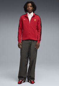 Rød lynlåsjakke med sorte sidepaneler, elastiske manchetter og et lille gult logo. Sammen med løstsiddende olivengrønne bukser og røde sneakers.