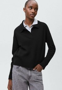 Čierny pletený sveter s golierom, s rebrovaným lemom a dlhými rukávmi, nosený cez bielu košeľu s golierom a skombinovaný so sivými džínsy.