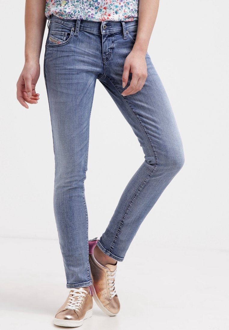 Personne portant un jean skinny bleu clair et des baskets lacées rose doré métallique, debout avec une jambe légèrement pliée.