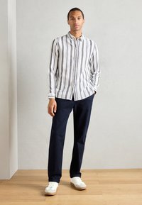 Striped button-up shirt in white, navy, and gray. Paired with navy pants and white sneakers. Slim fit with cuff details and neutral backdrop.