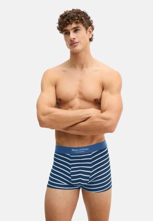 Homme torse nu aux cheveux bouclés, les bras croisés, portant un boxer rayé bleu et blanc de Marc O'Polo.