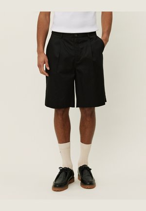 Pantaloni corti neri su misura con pieghe, realizzati in un tessuto liscio. Indossati con calze color crema e scarpe in pelle nera con suola in gomma.