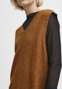 Kamizelka dzianinowa w ciepłym brązie, z dekoltem w serek i pionową fakturą, zestawiona z czarną, przezroczystą bluzką z długim rękawem i dekoltem z falowanym brzegiem.