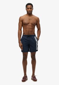 Shorts de bain bleu marine avec une taille à cordon et un grand logo blanc "SUPERDRY" sur le côté. Portés avec des chaussures marron.