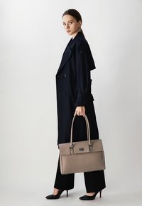 Svijetlosiva teksturirana torba s dvostrukim ručkama, uparena s tamnoplavim balonerom i crnim hlačama. Model stoji ispred neutralne pozadine.